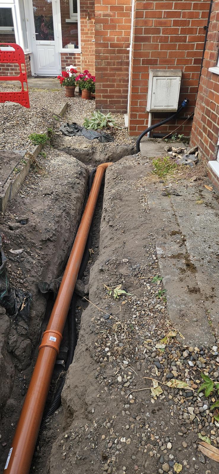 New orange drain pipe laid in an excavated trench alongside a house